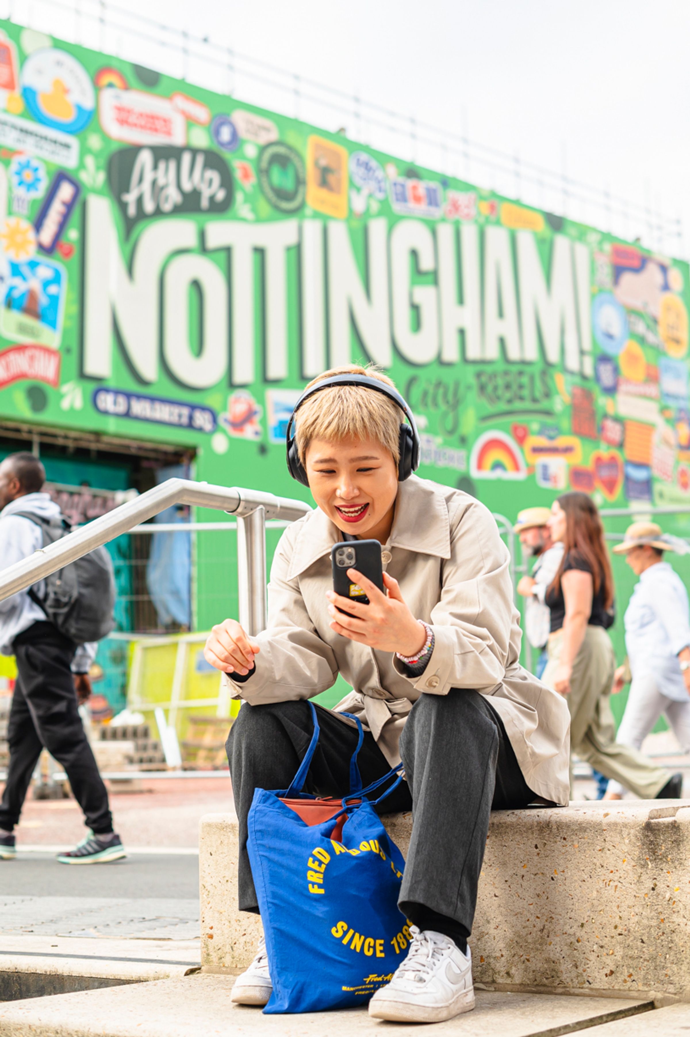 A woman wearing headphones smiles at her phone in front of a mural reading "Ay up, Nottingham!"