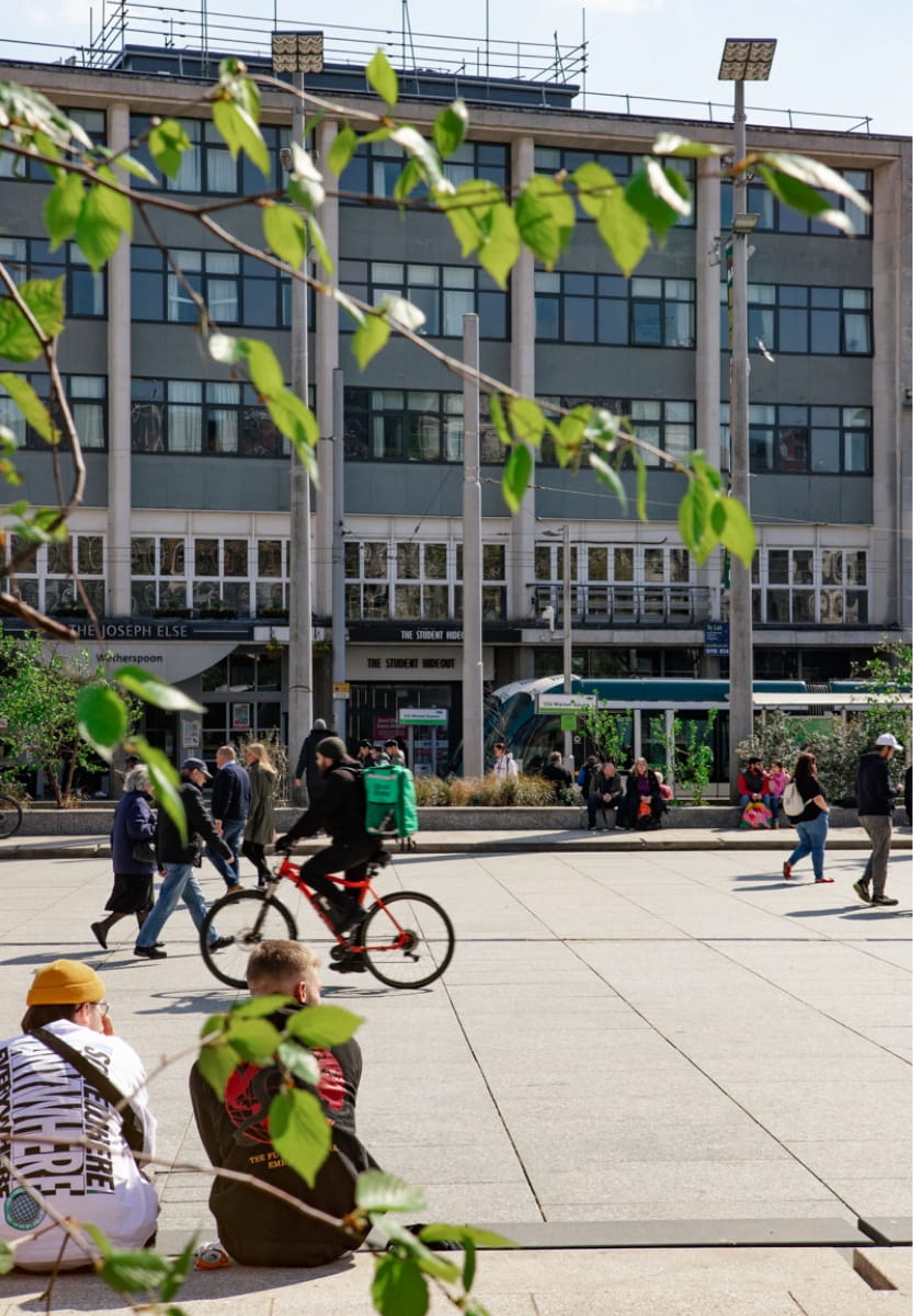 People walking and cycling across Old Market Square, Nottingham in the sunshine with tree branches in the foreground