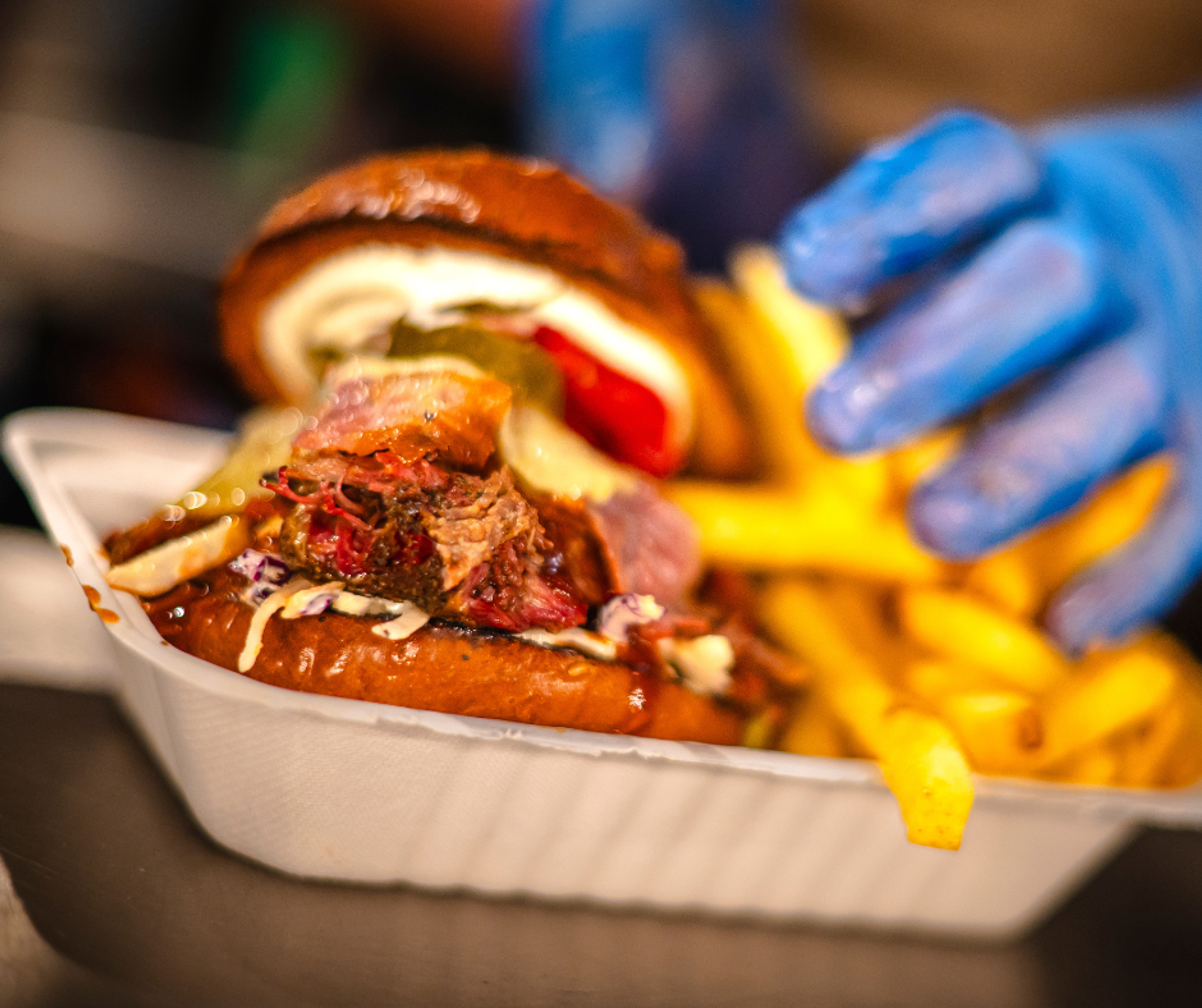 A dirty burger and chips being served in a takeaway container. The server is wearing blue catering gloves.