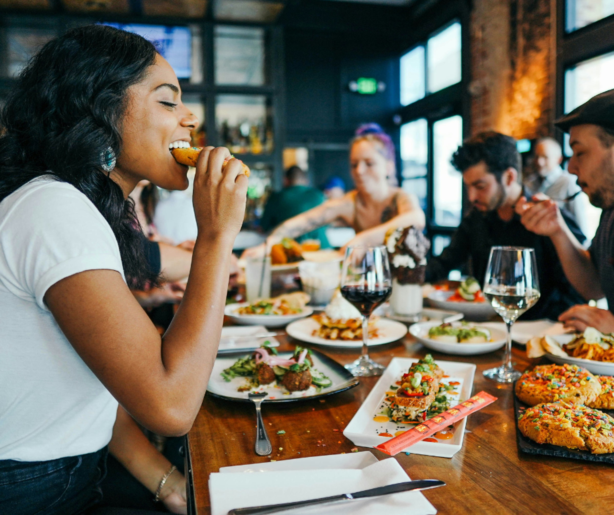 A woman biting into a cookie, in a restaurant with friends and a table of food and wine.