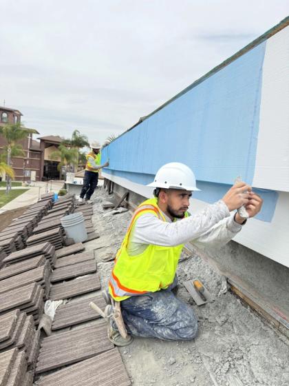 Crew member in hard hat and safety vest inspecting a trowel of base coat at a rooftop parapet, freshly coated blue section behind him