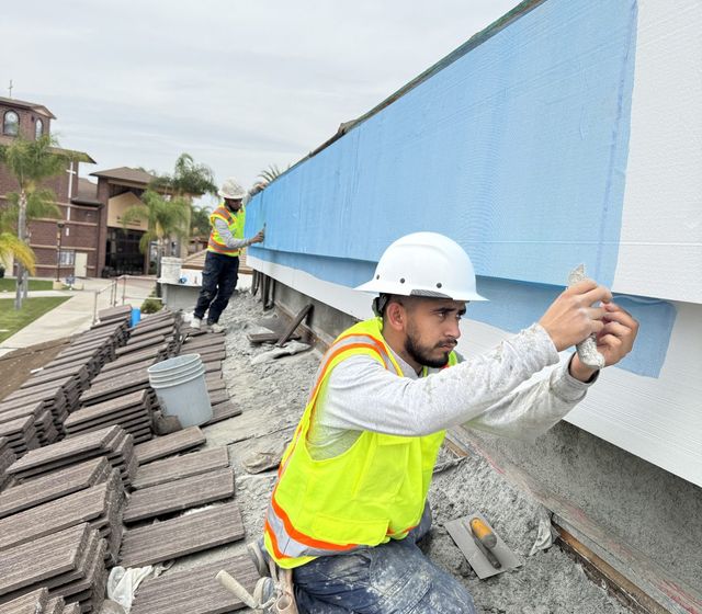 Crew member in hard hat and safety vest inspecting a trowel of base coat at a rooftop parapet, freshly coated blue section behind him
