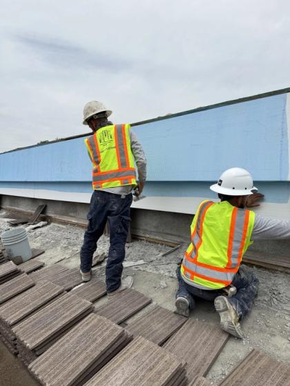 Two crew members in hard hats and hi-vis vests coating a long rooftop parapet band, roof tiles staged in the foreground
