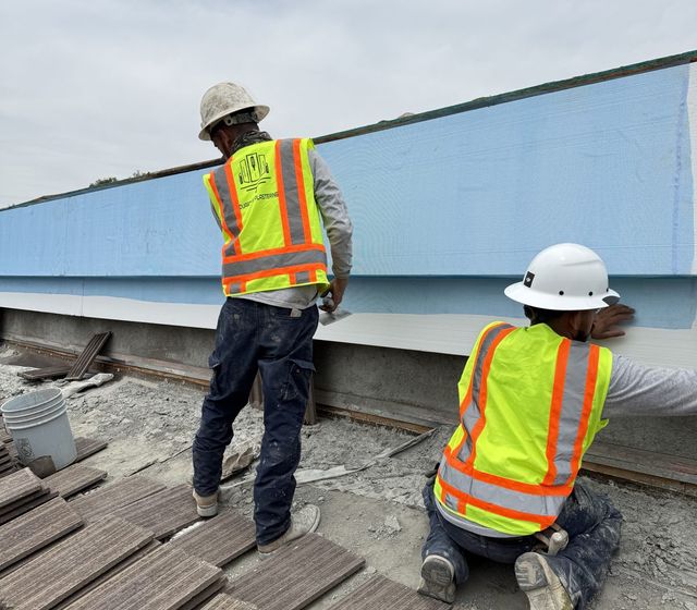 Two crew members in hard hats and hi-vis vests coating a long rooftop parapet band, roof tiles staged in the foreground
