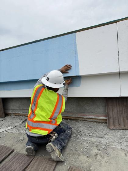 Plasterer kneeling on a roof deck, hand-checking the transition between base coat and finished white parapet