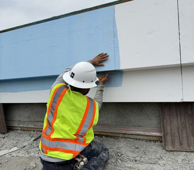 Plasterer kneeling on a roof deck, hand-checking the transition between base coat and finished white parapet