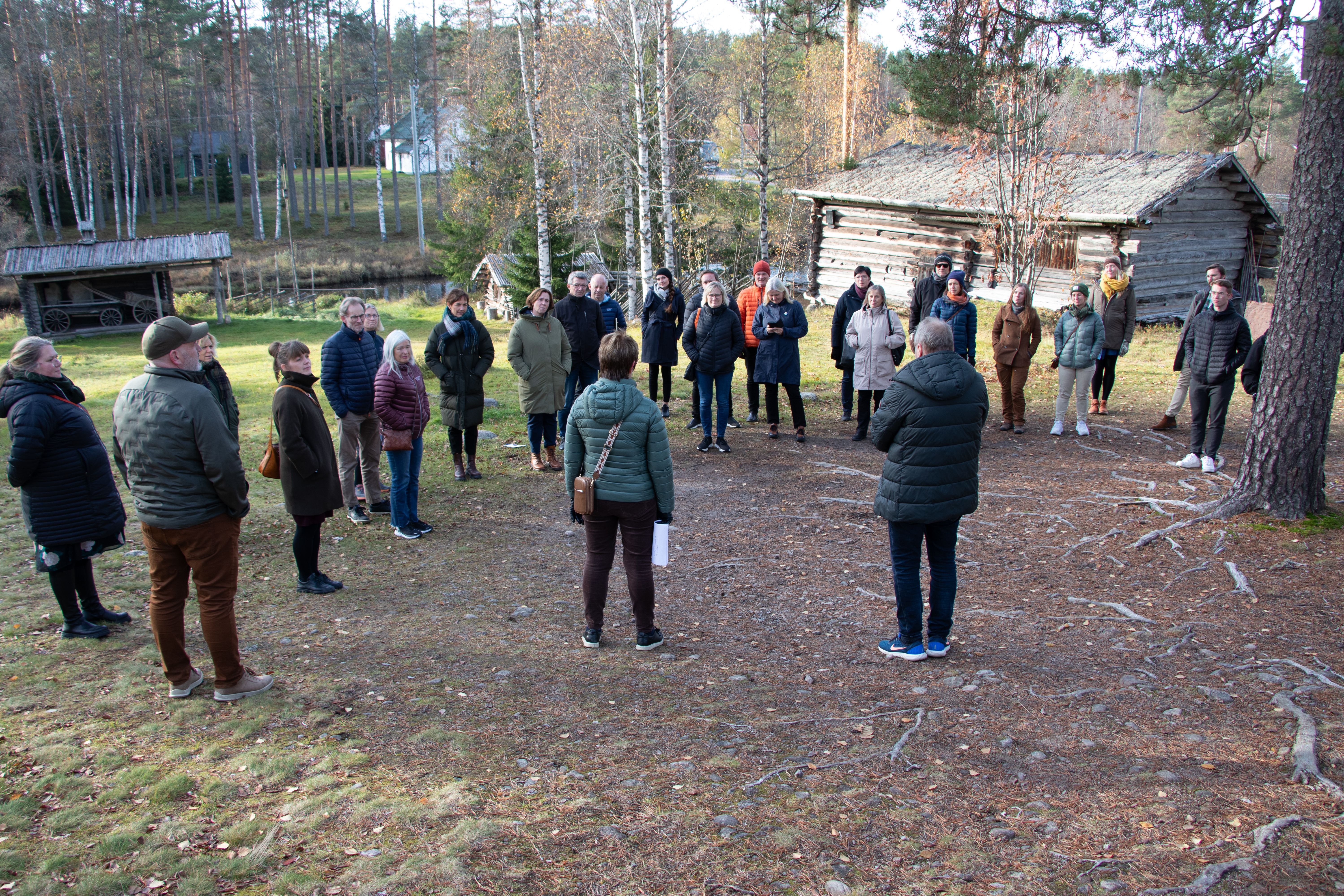 En gruppe mennesker som står i ring på en gressbakke under et furutre, foran en gammel tømret bygning