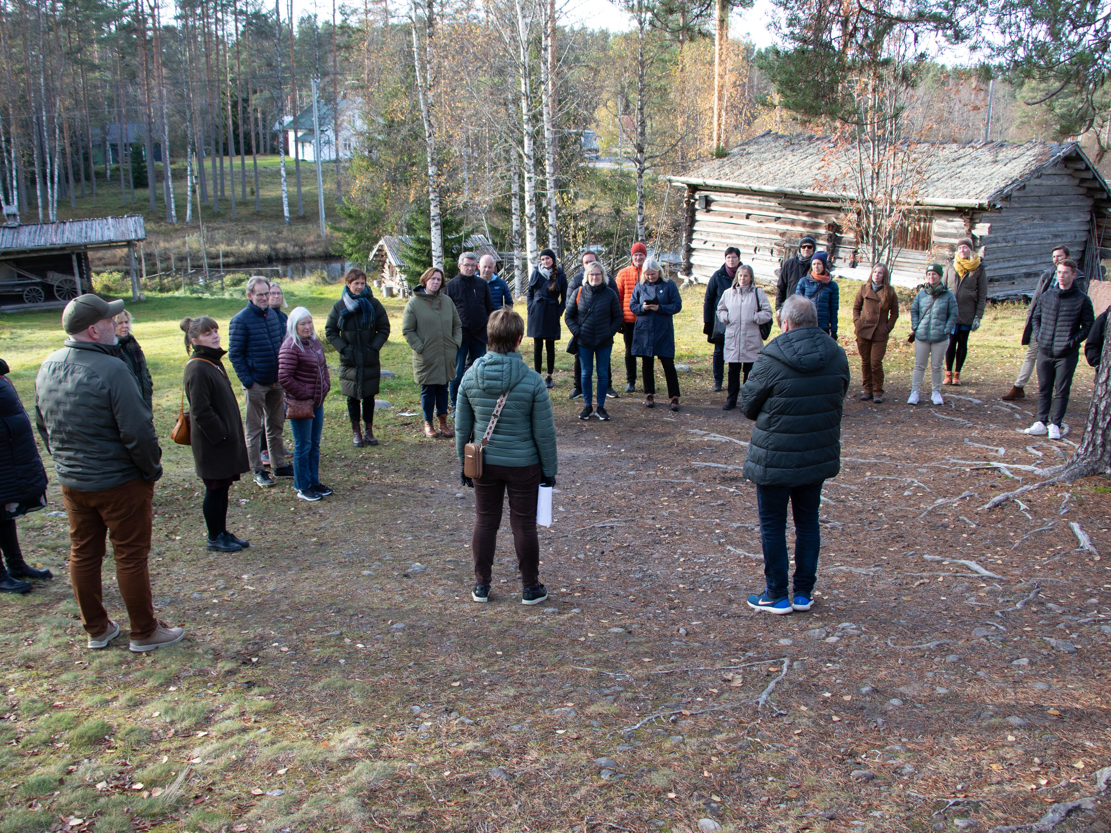 En gruppe mennesker som står i ring på en gressbakke under et furutre, foran en gammel tømret bygning