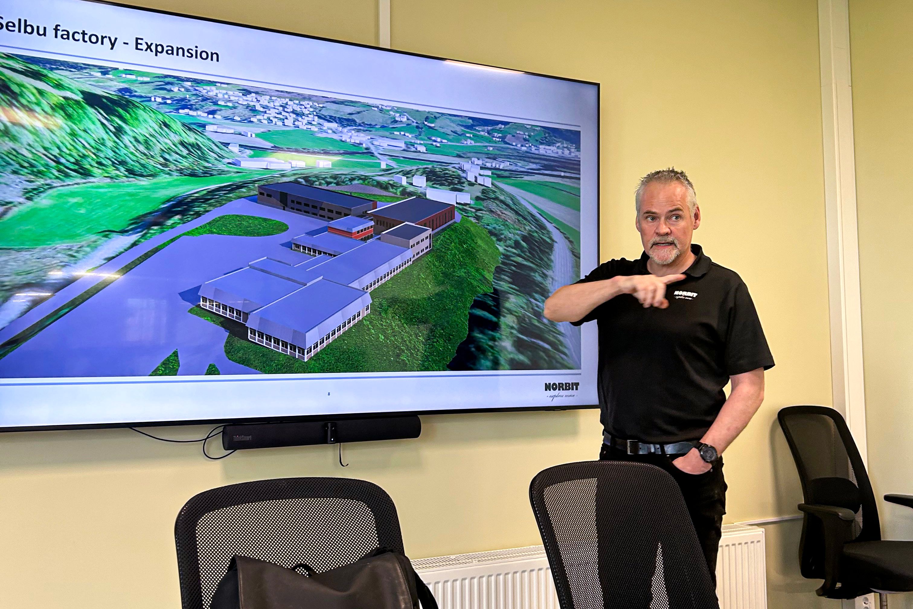 Mann presenterer, peker med høyre hånd. Skjerm med bilde av industriområde på veggen. 