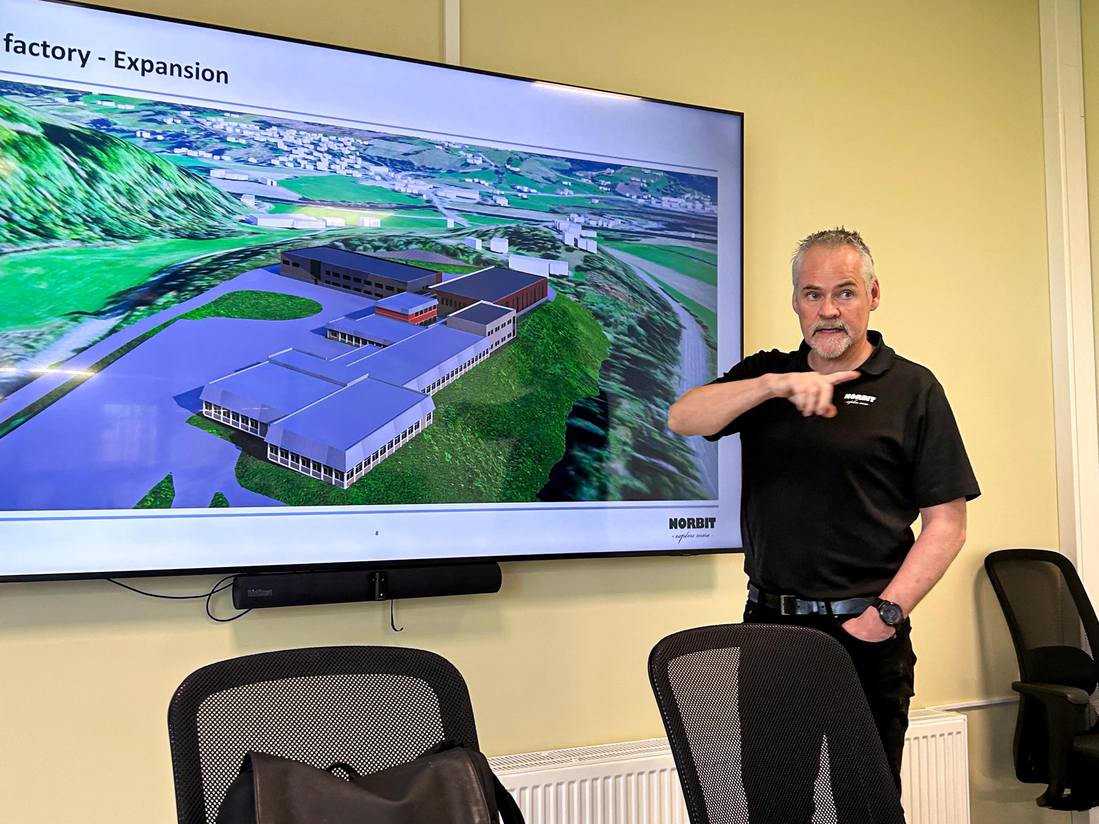 Mann presenterer, peker med høyre hånd. Skjerm med bilde av industriområde på veggen.