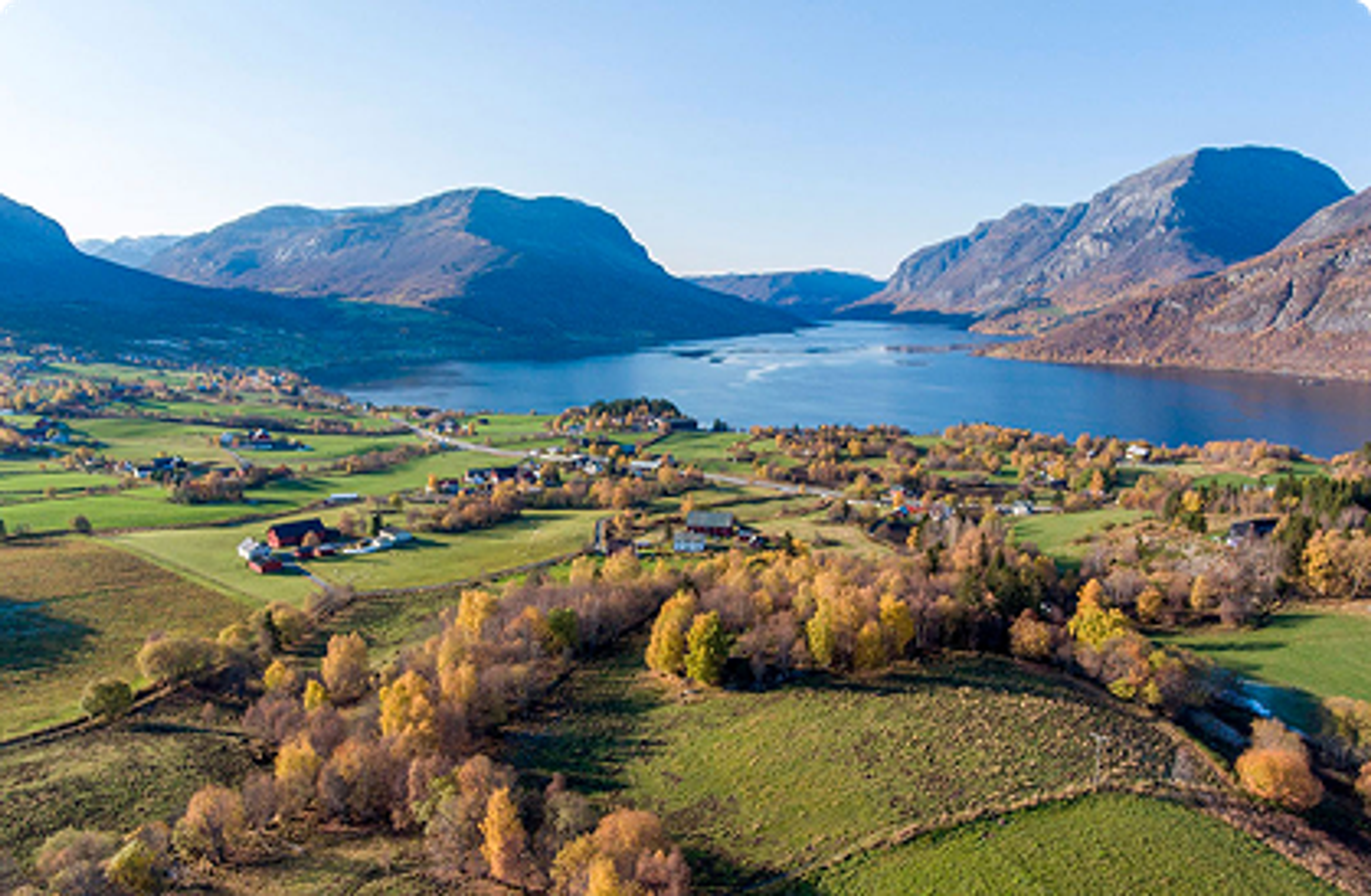 Landskapsbilete med ei grend, ein fjord og fjell bakom. Haustfargar.