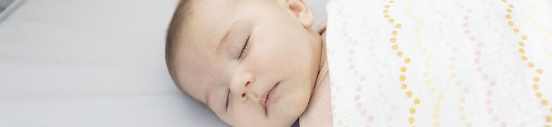Un petit bébé endormi sur un matelas, emmailloté dans une couverture blanche et colorée.