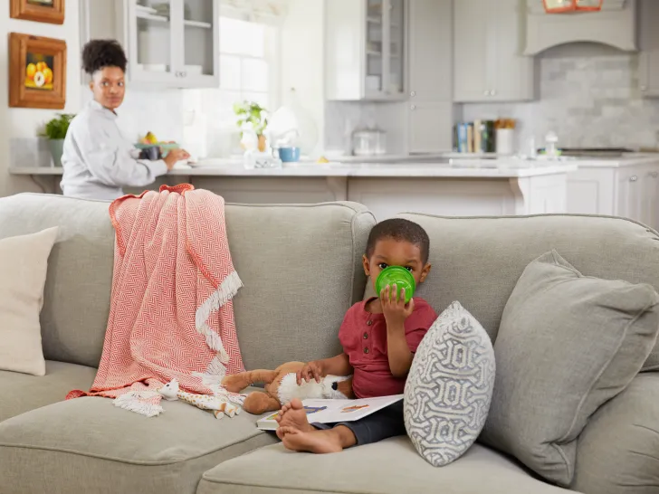 A young boy sat on a couch drinking from a beaker with a book and teddy on his lap with a woman in the background watching over him.