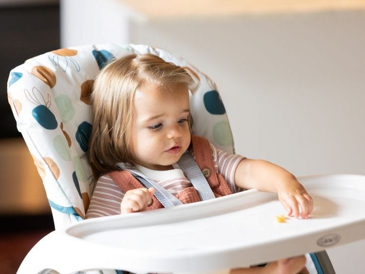 A young girl sat in a high chair looking at the fruit on the tray that she is picking up.