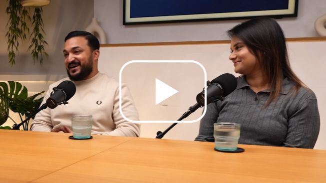 A couple sat at a table with Microphones. Both are looking away from the camera, the man is talking. A white play button is over the top.