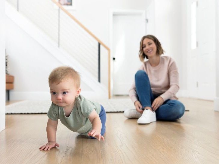 A young baby crawling along a wooden floor with a woman sitting in the background watching.