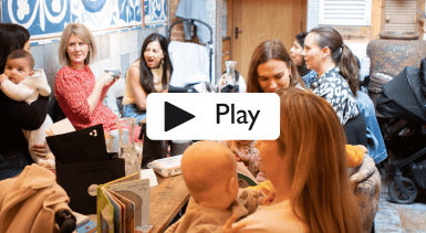 A group of mums sitting round a table talking, some are holding babies with books, wipes and bags on the table between them. With the word Play over the top of the image.