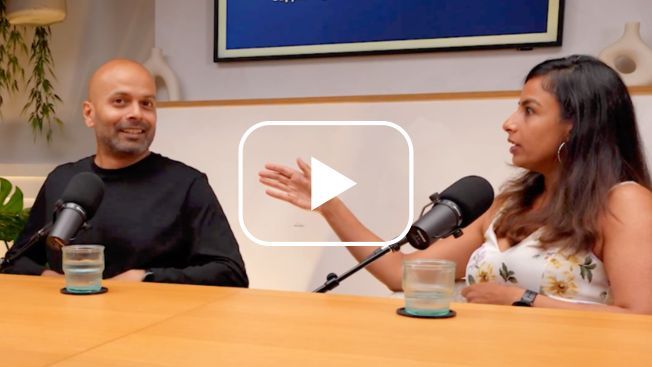A couple sat at a table with Microphones. The man is looking at the camera and the woman is looking away talking. A white play button is over the top.