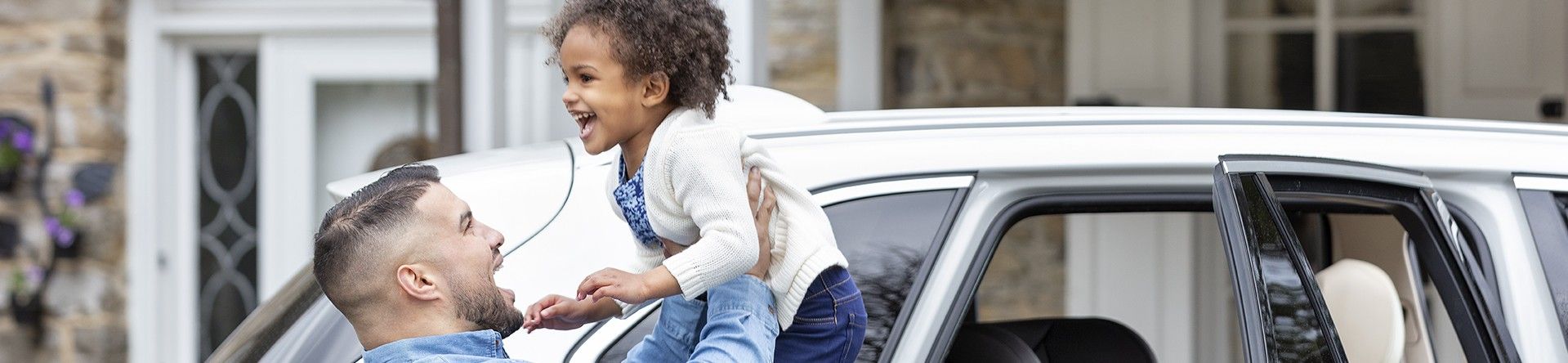 A man holding a young girl in the air in front of a white car with an open door.