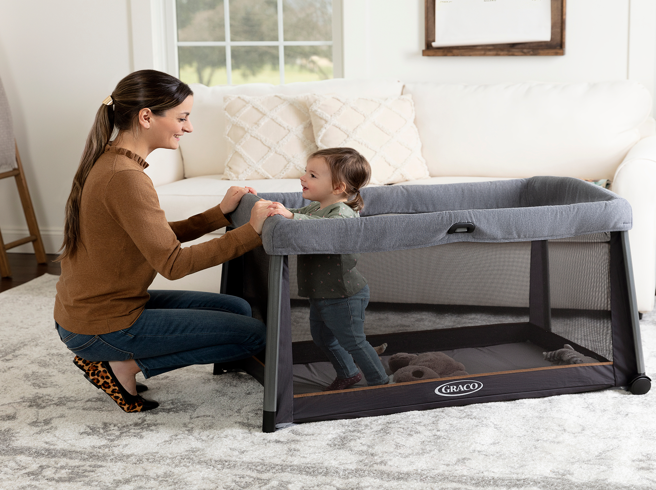 A toddler standing in travel cot beside her mum.