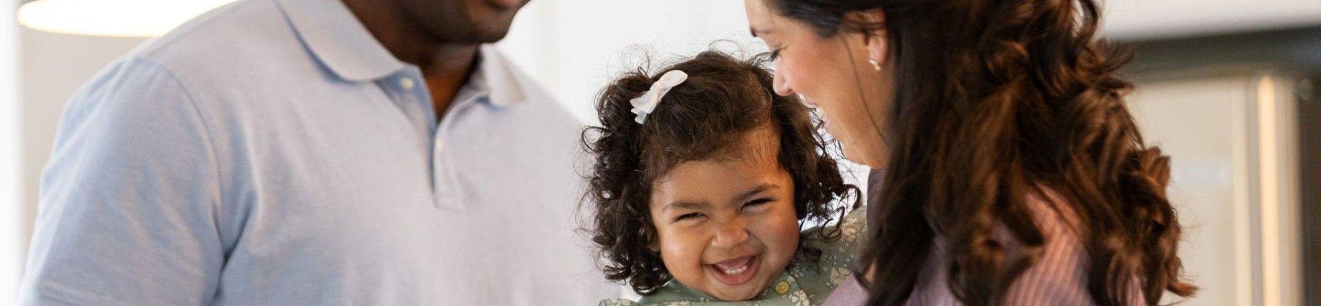 Ein lächelnder Mann steht hinter einer Frau, die ein lächelndes kleines Mädchen hält.