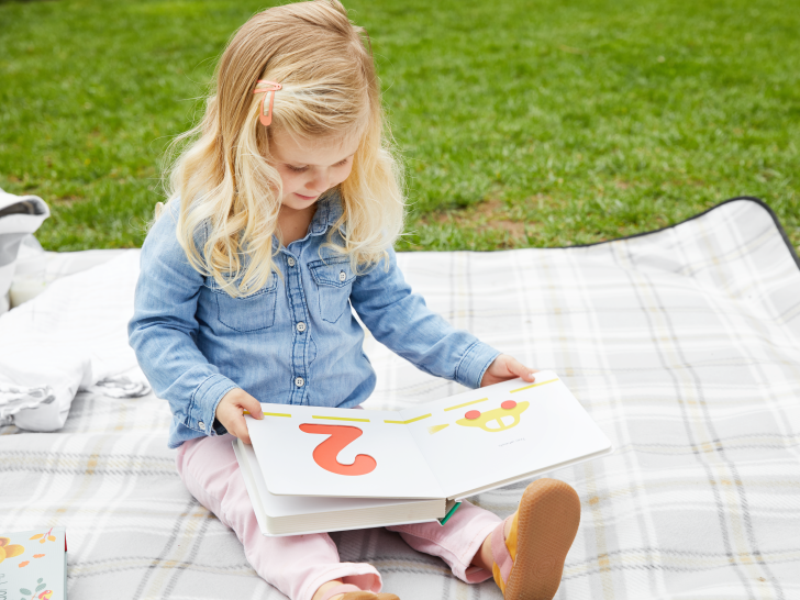 Ein junges Mädchen sitzt auf einer Decke im Gras und liest ein Buch.