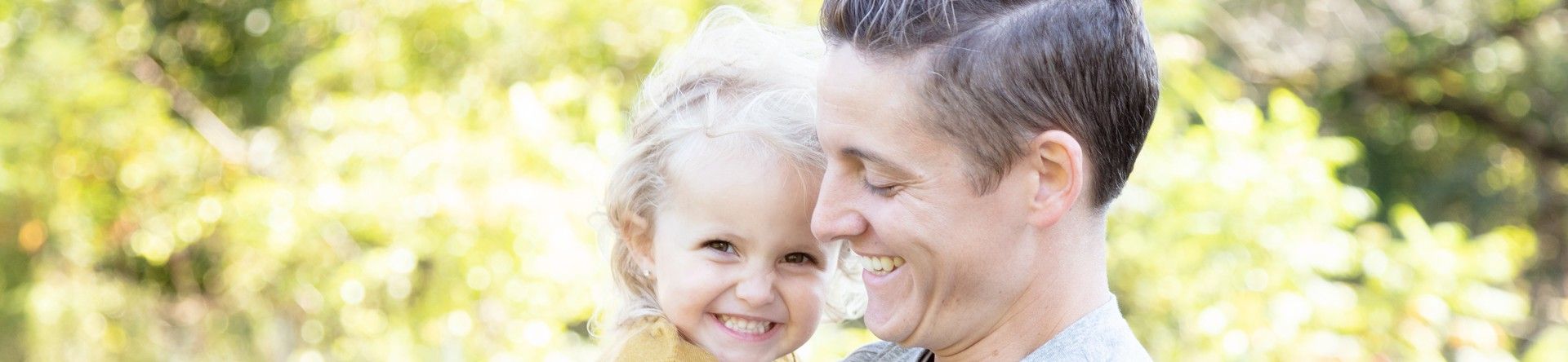 A man is staring outside holding a toddler girl smiling at her while she is smiling at the camera.