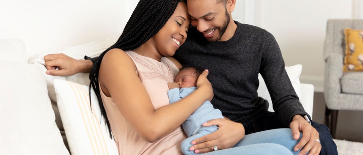 A mum sat on the sofa cradling a sleeping baby leaning into the dad sat next to them.