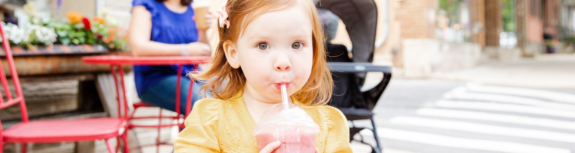 Ein junges Mädchen trinkt draußen einen Smoothie, im Hintergrund sitzt eine Frau an einem Tisch.