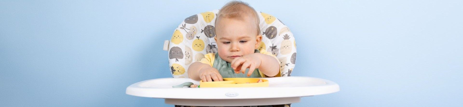 A young boy sitting in a highchair looking at the food on the tray grabbing it with both hands.