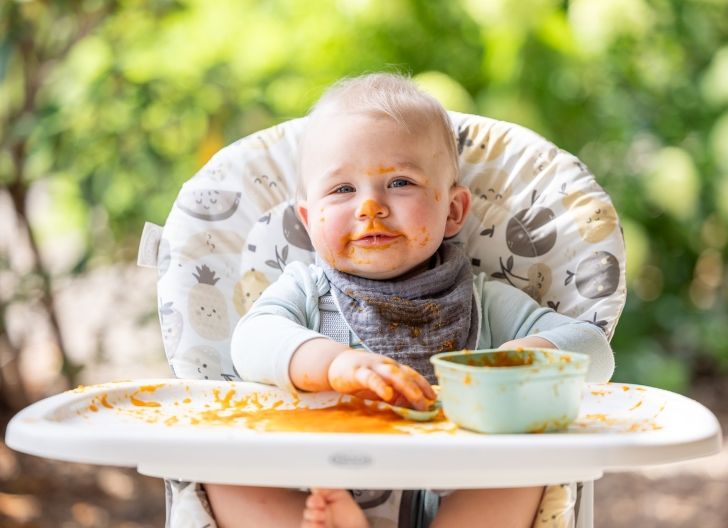 Ein Kleinkind sitzt im Hochstuhl mit Essen im ganzen Gesicht und auf dem Stuhl