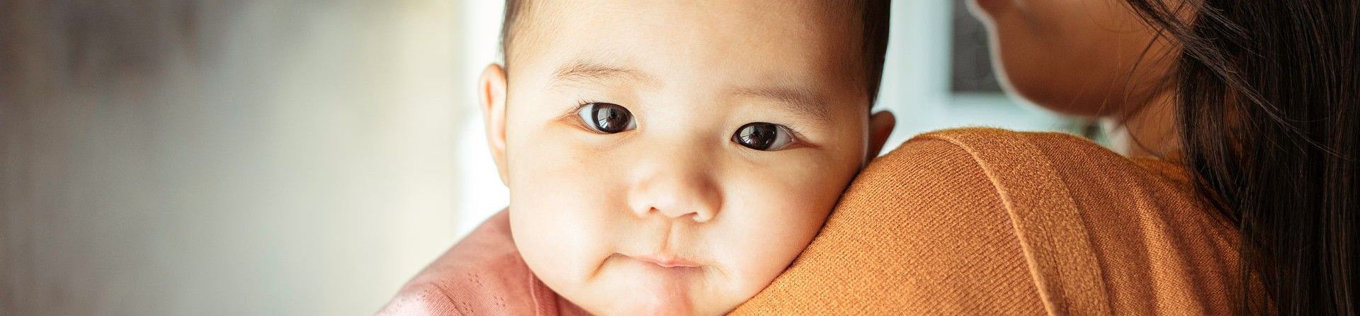 A young baby looking into the camera, being held by a woman with her back to the camera.