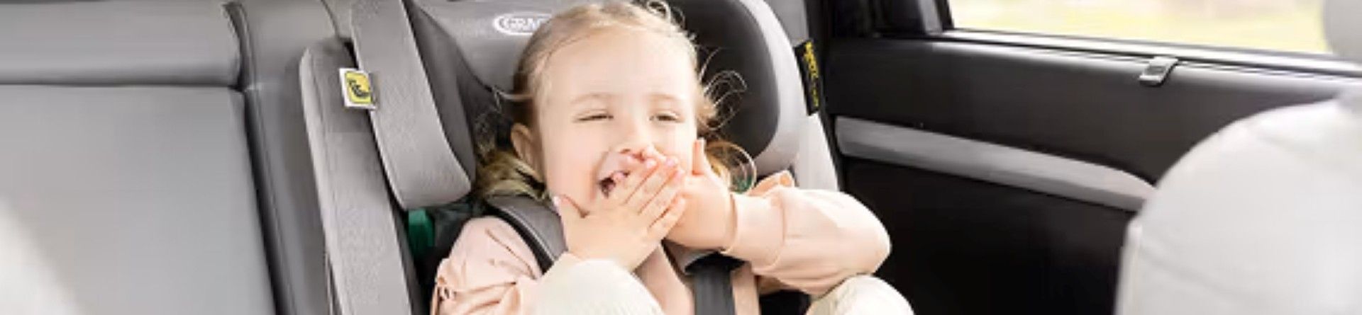 A girl sat in the back of a car in a car seat with her legs up in the air and her hands covering her mouth.