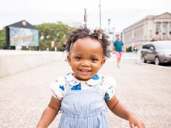 A young girl walking outside along a pavement, smiling at the camera.