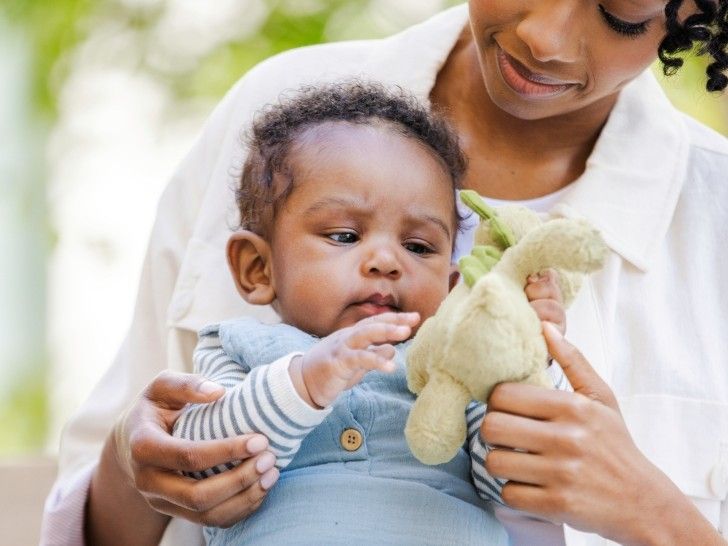 A young toddler sitting on a woman's lap holding and looking at a green soft toy