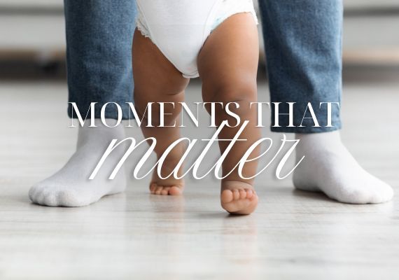 A close up of a baby with bare legs walking with an adults feet either side. The words Moments that matter is written over.