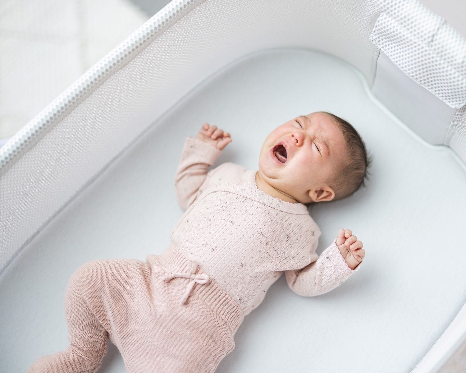 A young baby lying on her back in a cot crying.