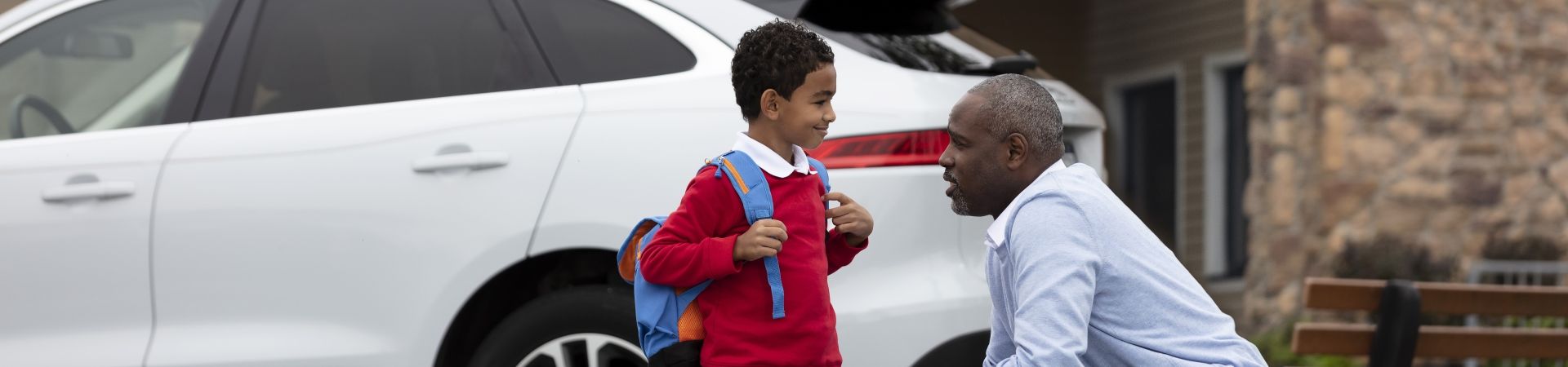 Ein Junge in Schuluniform mit Rucksack steht vor einem Auto, daneben hockt ein Mann