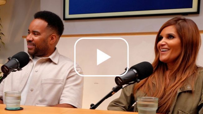 A couple sat at a table with Microphones. Both are looking away from the camera and smiling. A white play button is over the top.