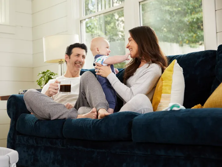 A man and woman sat together on a couch, the man is holding a cup of coffee looking at the woman who is holding a baby.