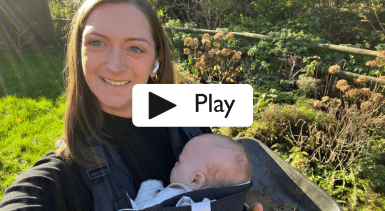 A woman outside in the countryside smiling at the camera holding a baby in a baby carrier.