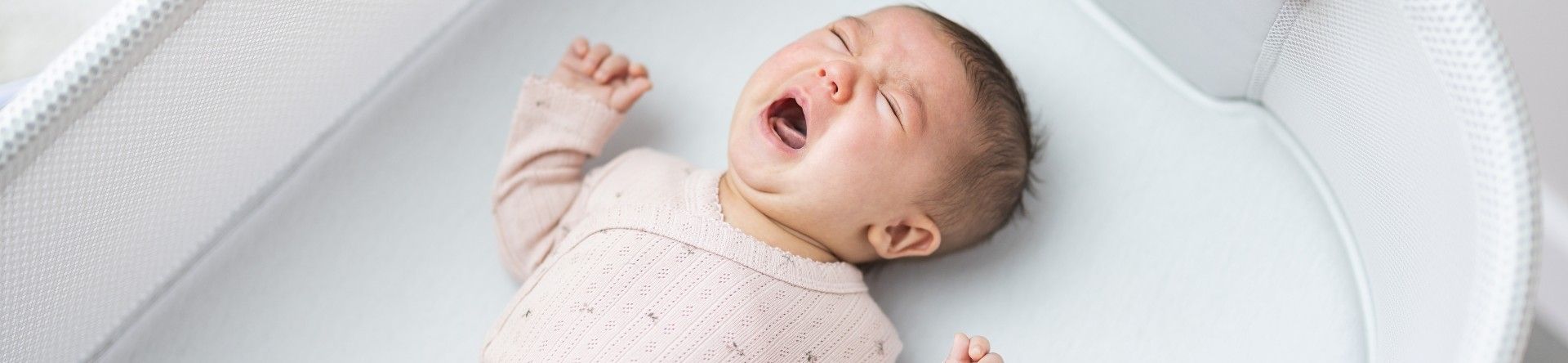 A young baby lying on her back in a cot crying.