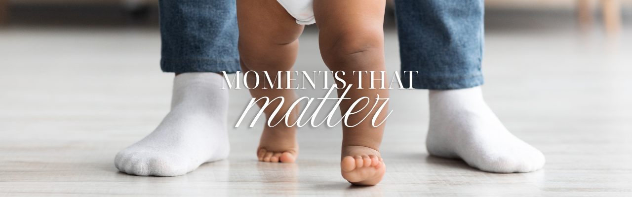 A close up of a baby with bare legs walking with an adults feet either side. The words Moments that matter is written over 
