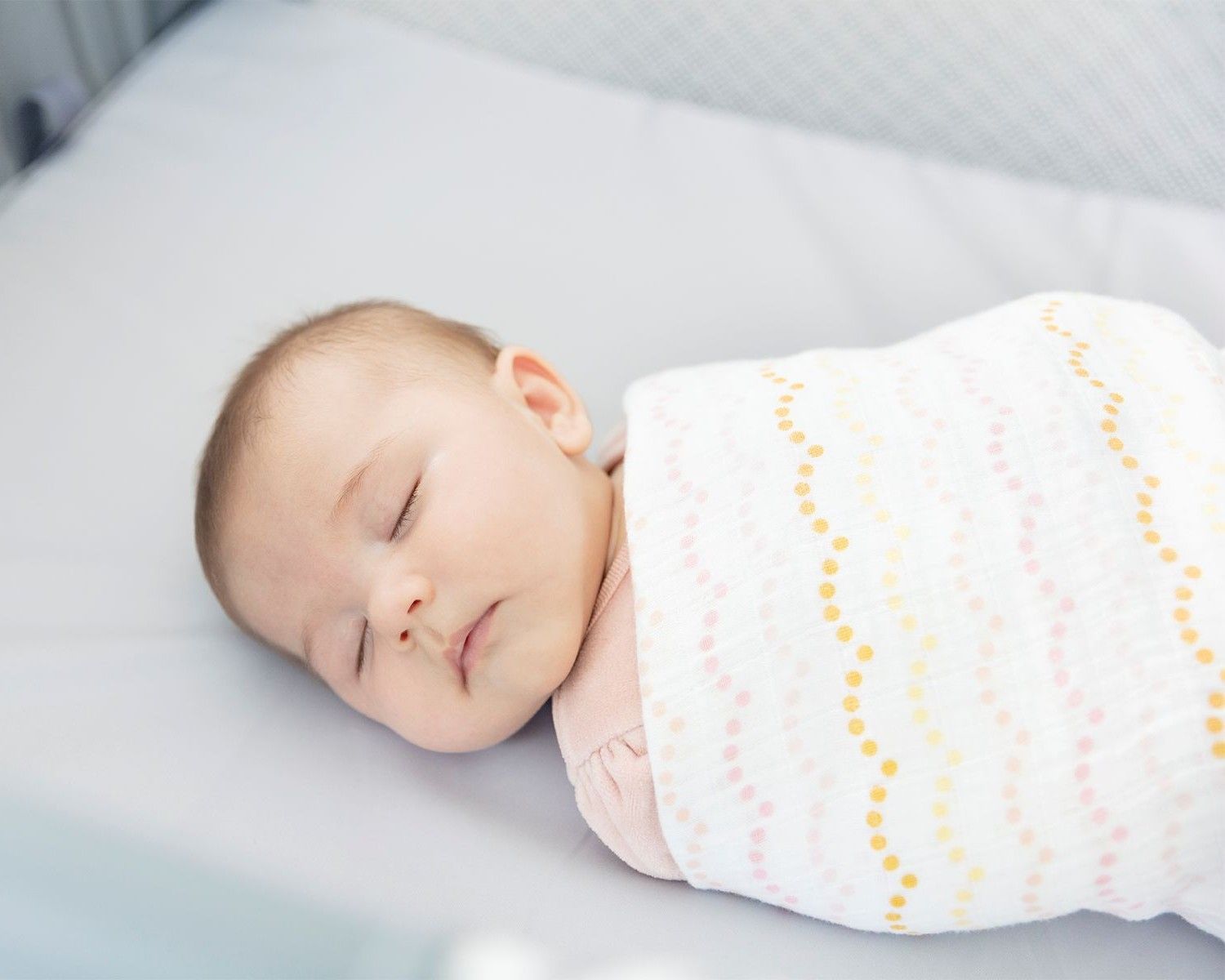 Un petit bébé endormi sur un matelas, emmailloté dans une couverture blanche et colorée.