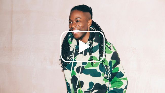 A man is standing against a cream background, looking away from the camera, smiling and wearing a green patterned hoodie. A white play button is over the top.