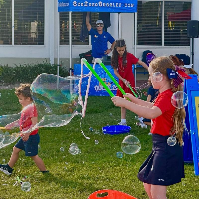 two kids with tri-string wands making a giant bubble beside Big Idea Bubbles signs