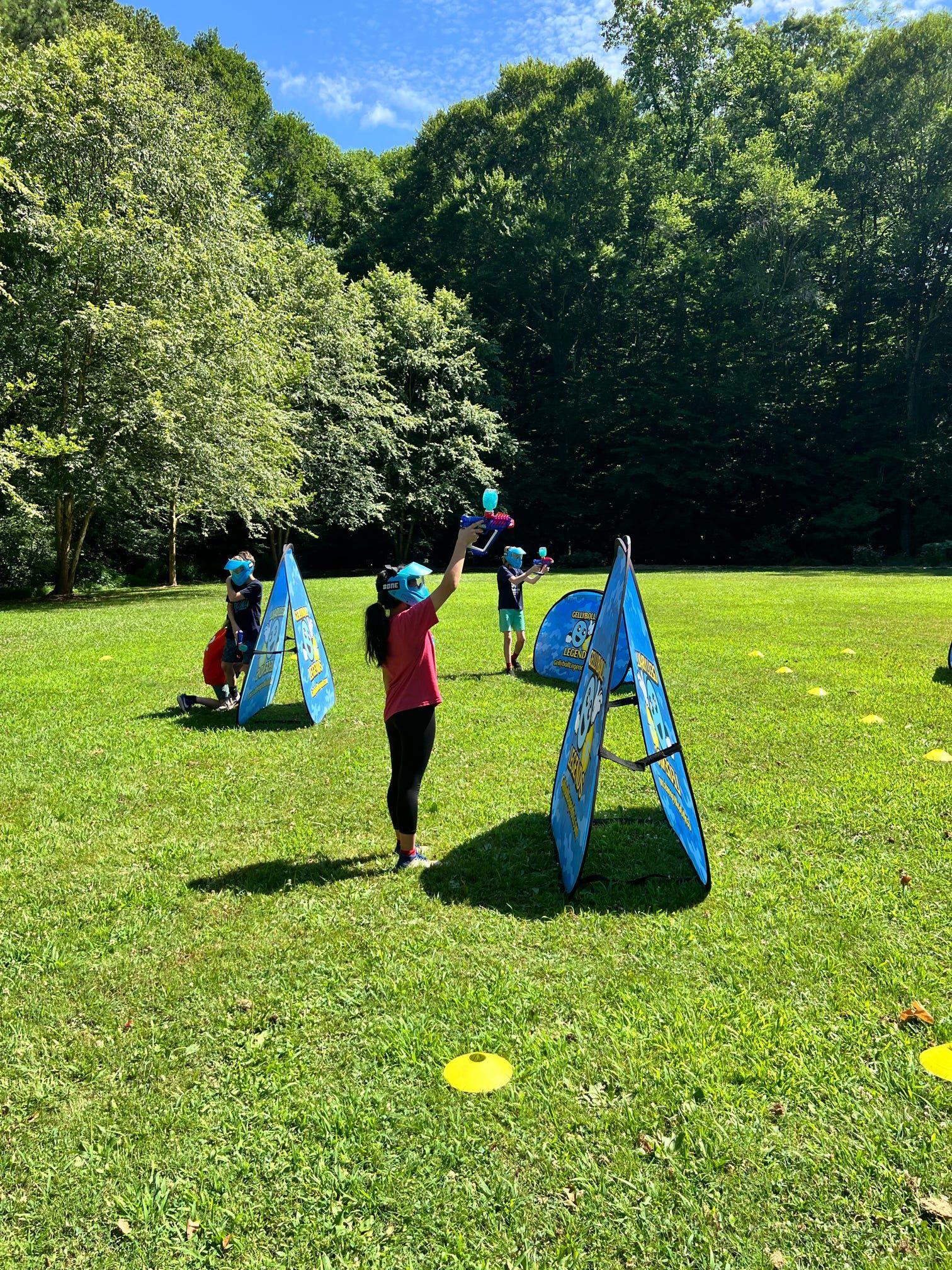 A girl in a blue mask raises her arm between bunkers as kids play across a park field