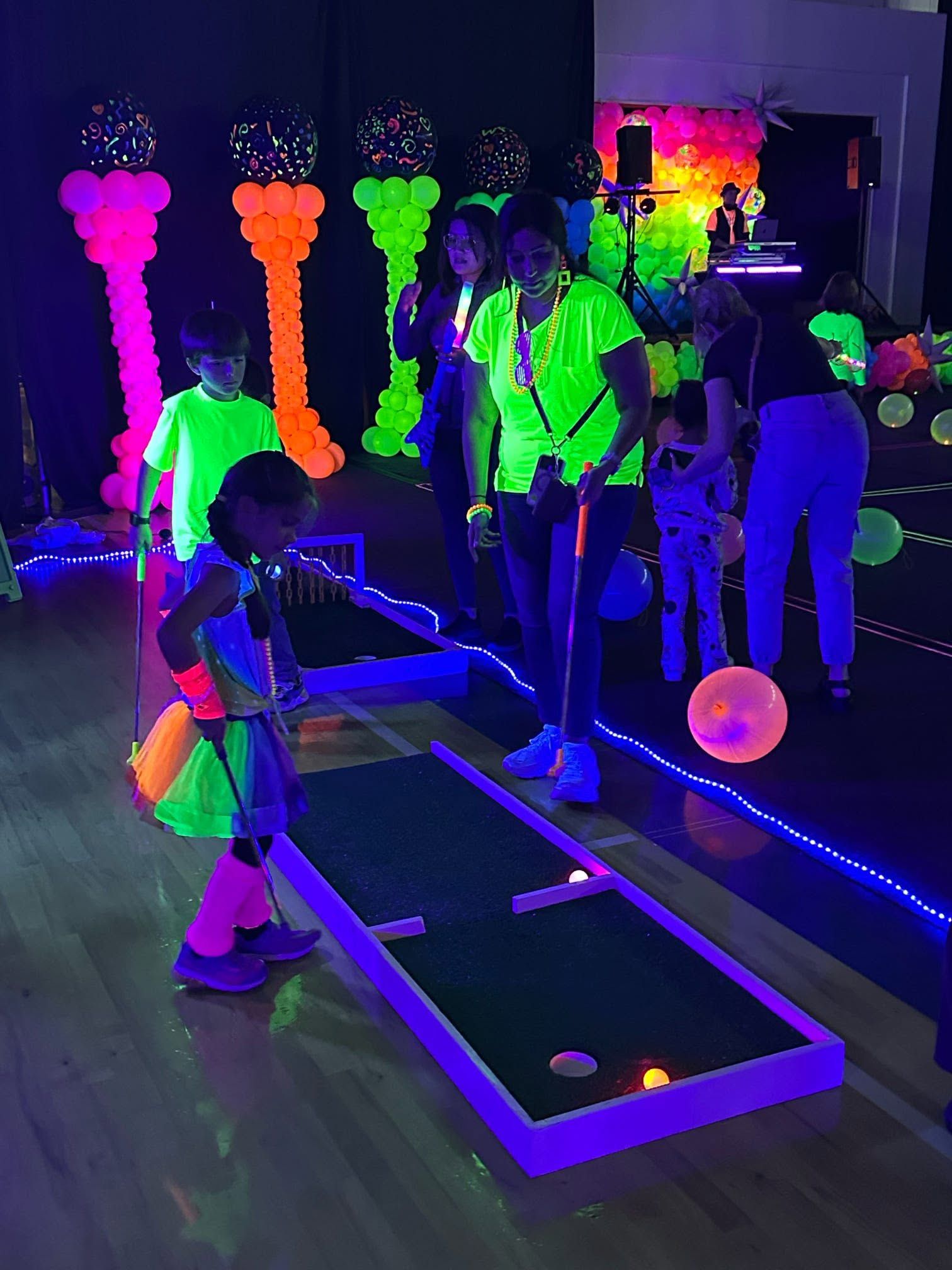 children playing a blacklit mini golf hole in a dark room with glowing neon obstacles