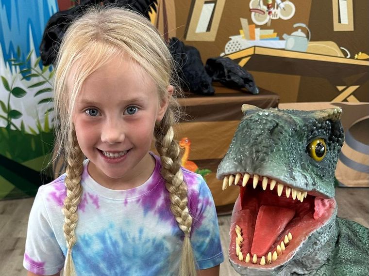 A girl in a tie-dye shirt smiles next to a lifelike baby T. rex