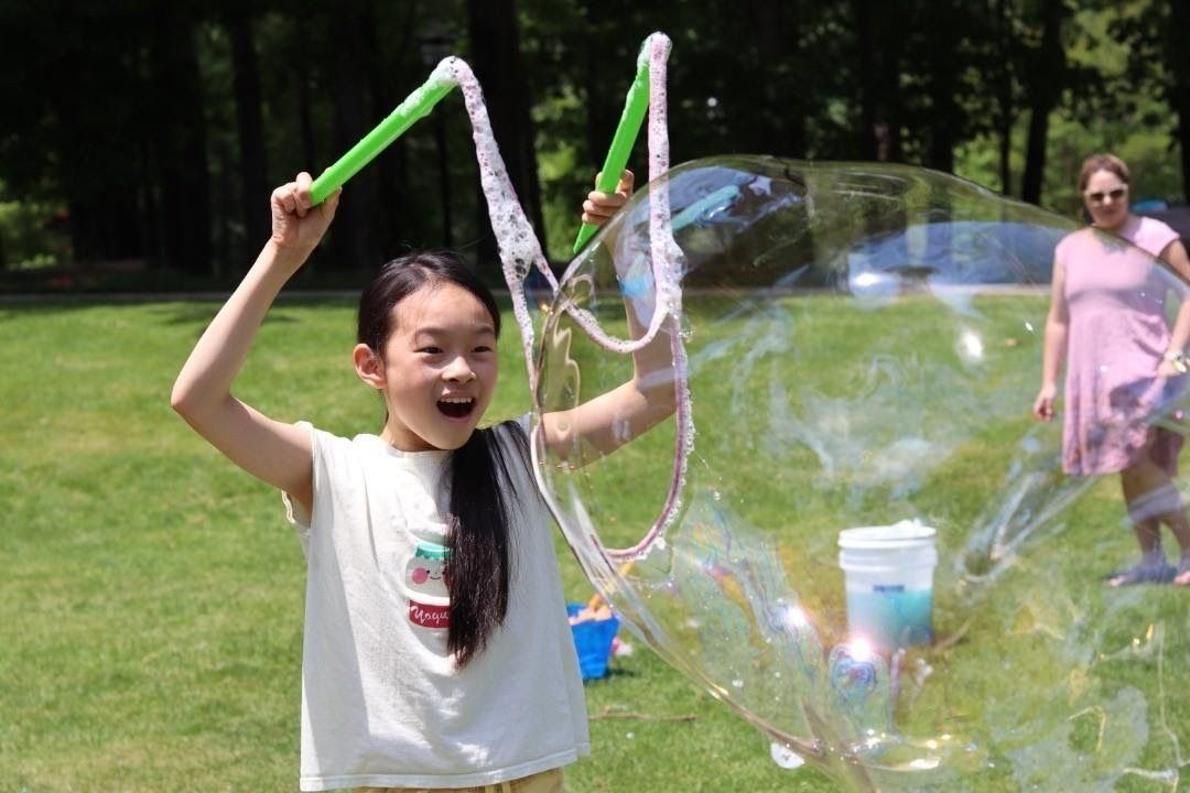 a girl with two wands raised making a giant bubble in a backyard